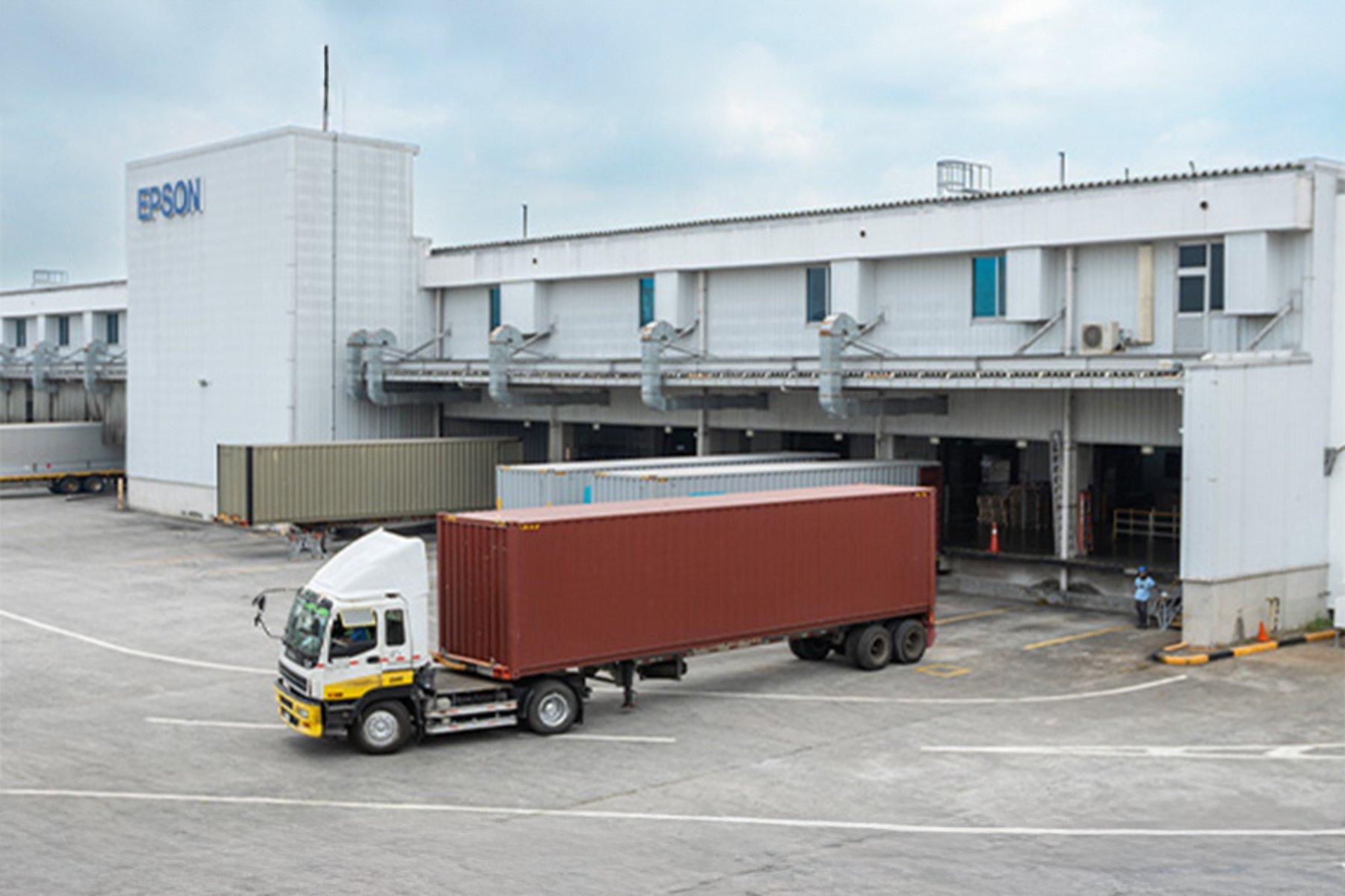 A large truck with a red shipping container is parked in front of a white industrial building with "Epson" signage. The scene is calm and orderly.