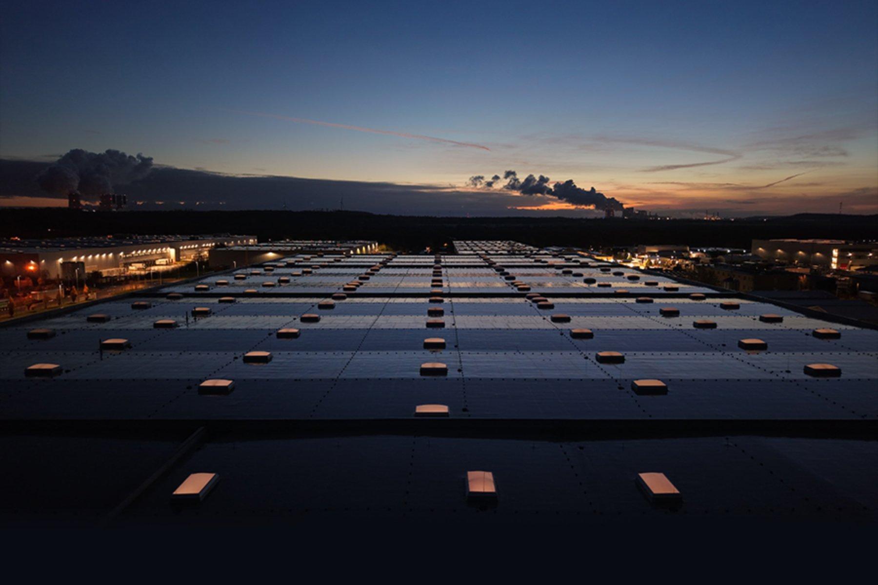 Rooftop view of an industrial building at dusk, with a grid of skylights glowing warmly. Smokestacks and a vibrant sunset silhouette the horizon.