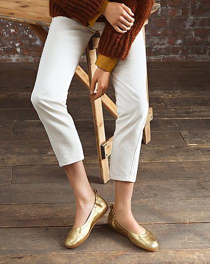 A woman wearing white trousers and gold ballerina shoes.