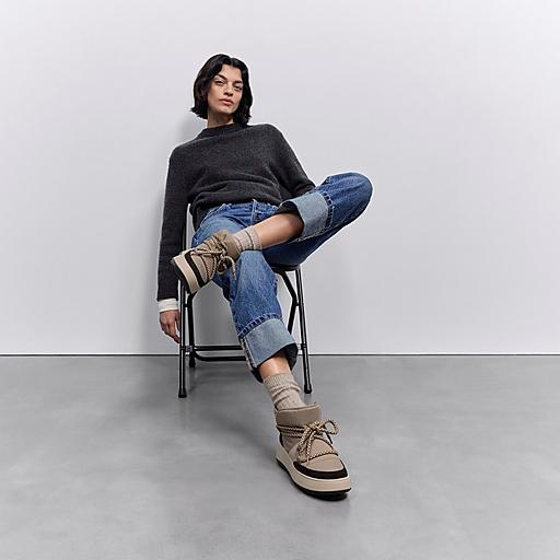 Persona sentada, con las piernas cruzadas, vestida con un suéter gris, pantalones vaqueros remangados y zapatillas deportivas color canela con detalles de cuerda.