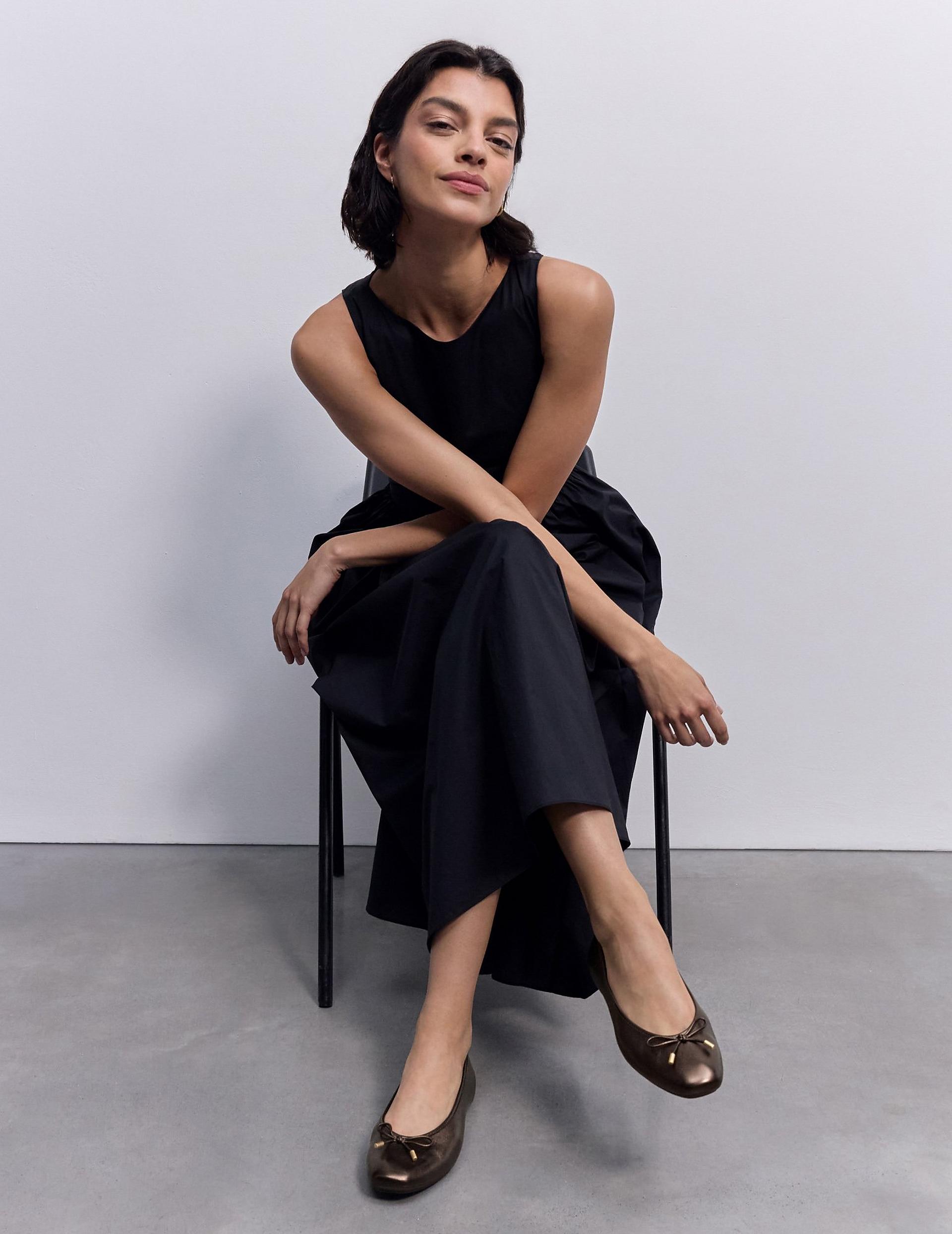 Model seated on a chair wearing a sleeveless black jumpsuit and metallic bronze ballet flats.