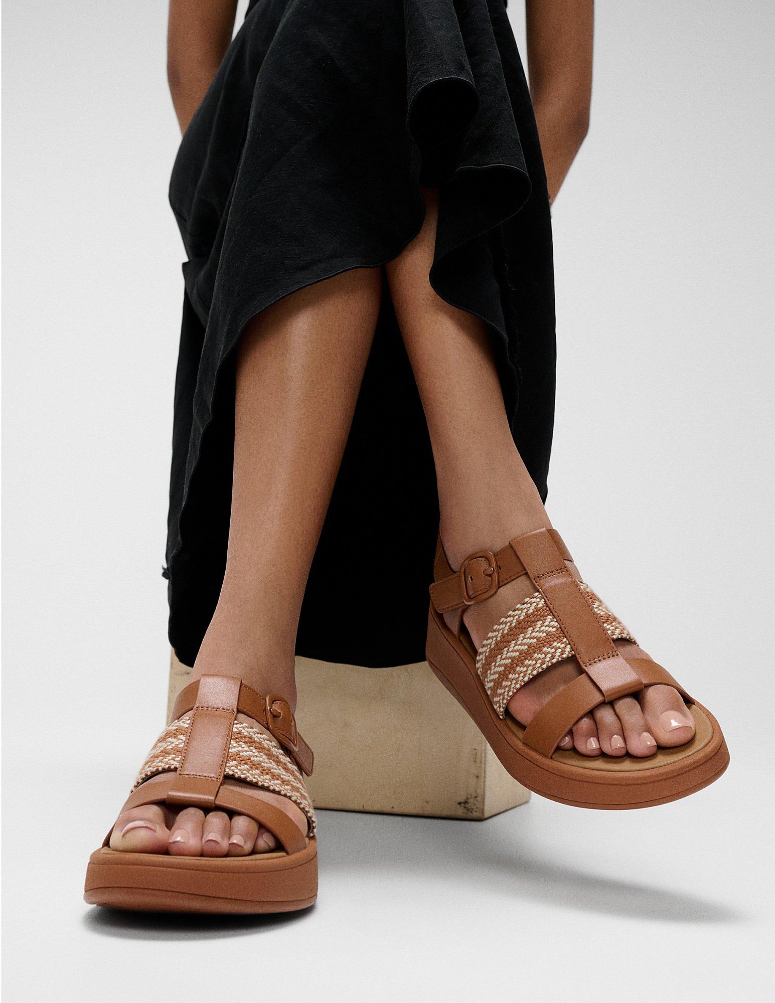 Woman sitting on a stool wearing black skirt and FMODE weave sandals in Tan - From FitFlop US