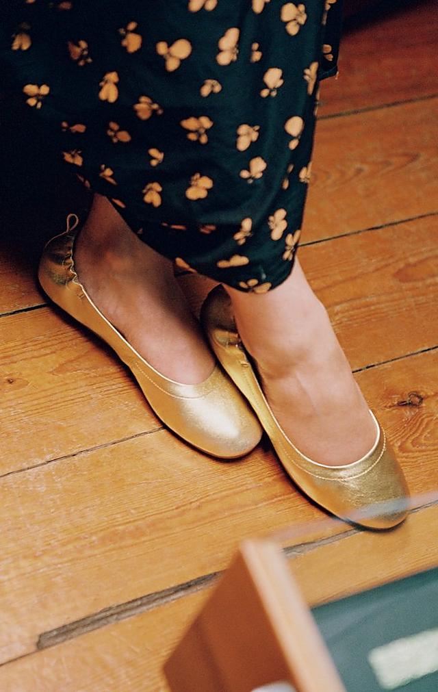 Woman wearing a black floral dress is modelling ballerina shoes in gold.