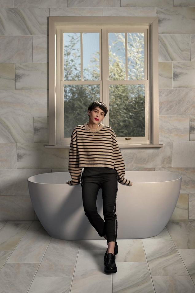 Woman sitting on bathtub edge in bathroom with white marble tile floors and walls.