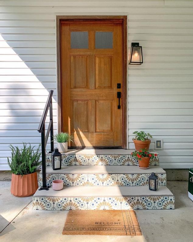 A porch with a light wood front door, patterned tile stairs, and several plants. 