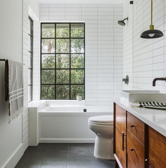 A bathroom with slate-colored floors, white wall tile, a chestnut-colored vanity, and large windows.