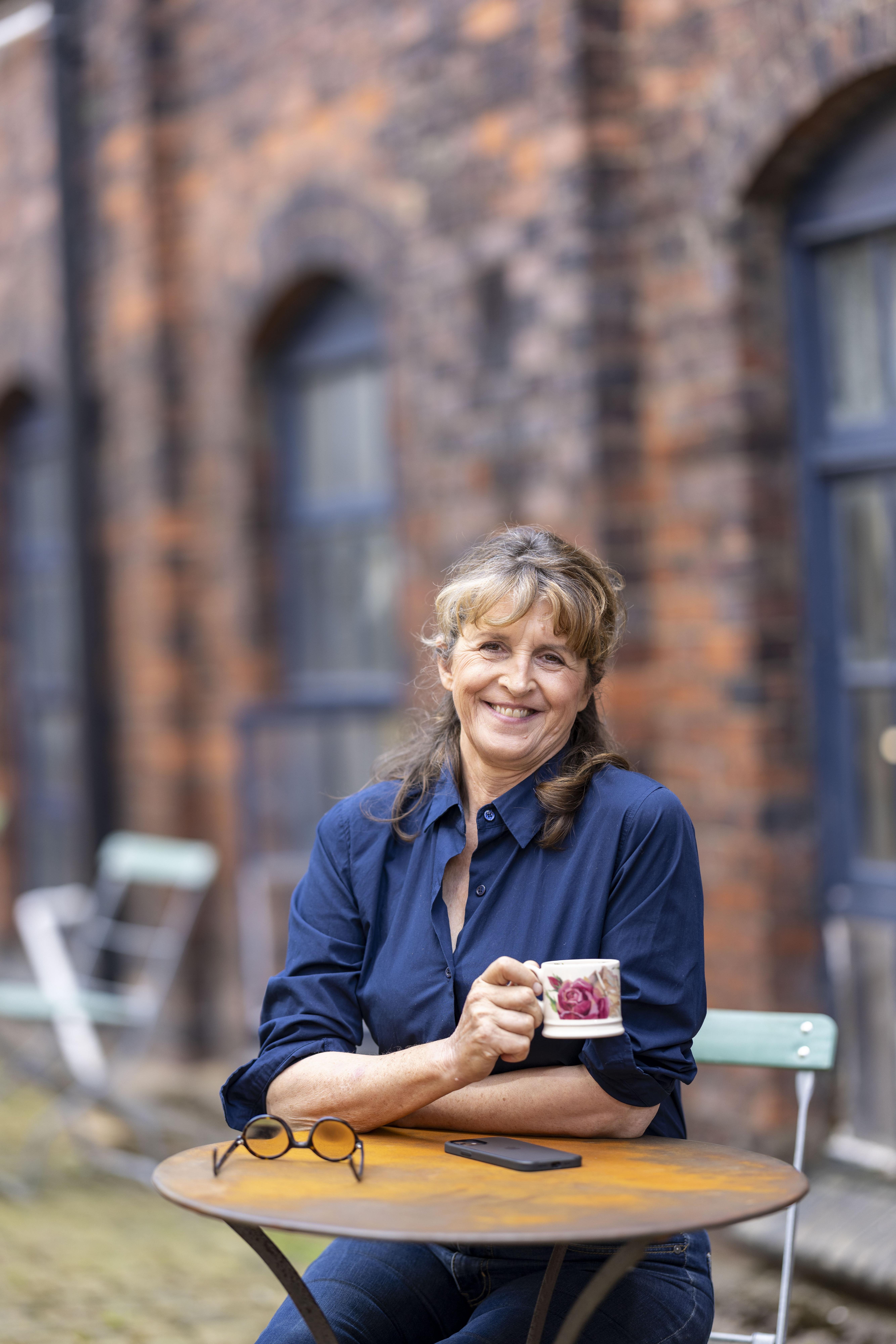 Emma Bridewater sits at a table in front of a redbrick factory, she holds a mug with a floral design