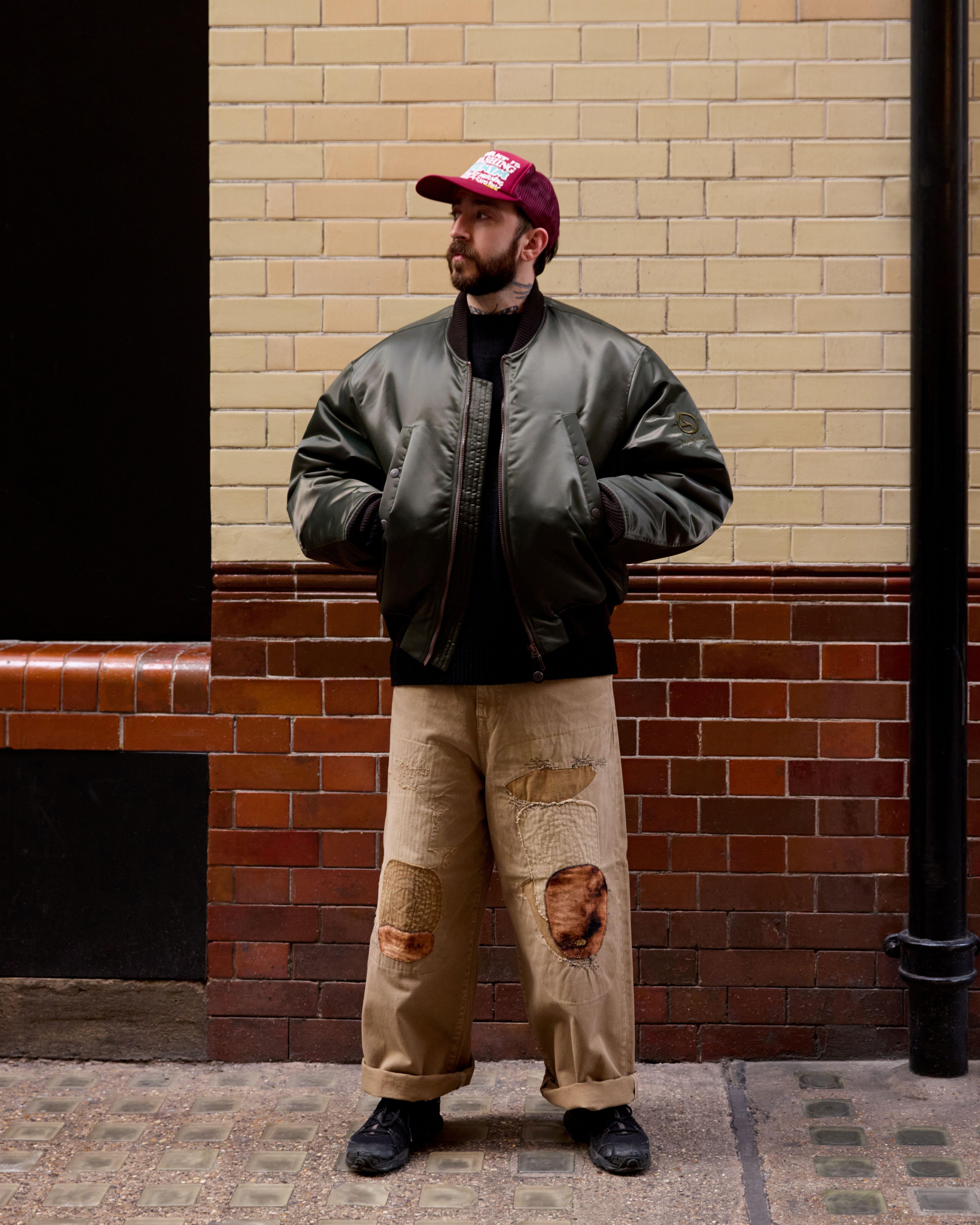 A man stands against a brick wall wearing a red baseball cap, MA-1 jacket and embellished jeans