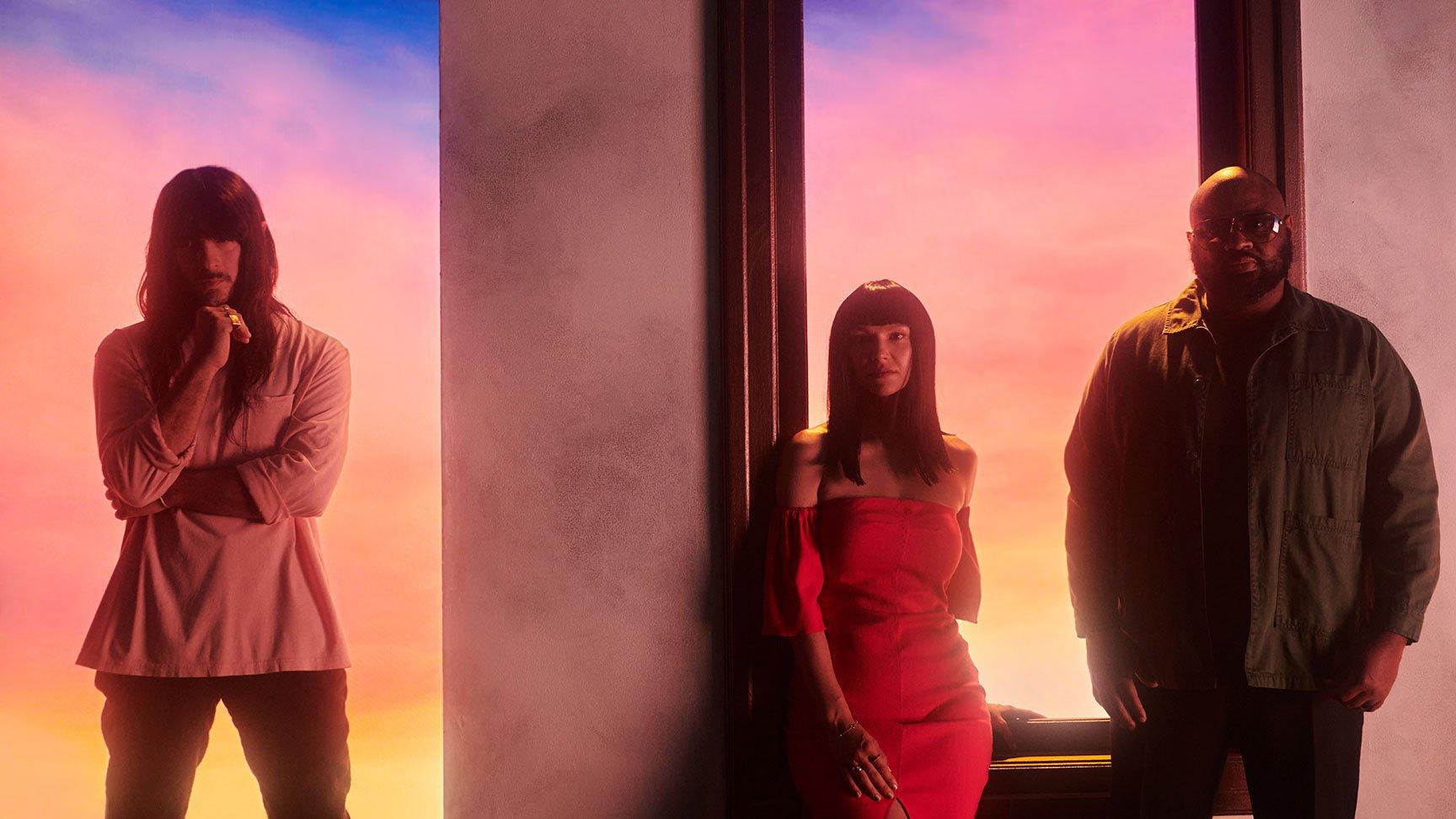 Khruangbin poses against a softly backlit pastel background featuring a sunset