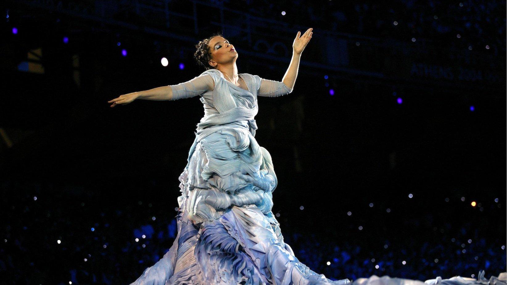 Photo of Bjork performing on stage at the opening of the 2004 Athens Olympics. Bjork is wearing a lavish dress with multiple shades of blue. Her hands are in an L shape in the air.