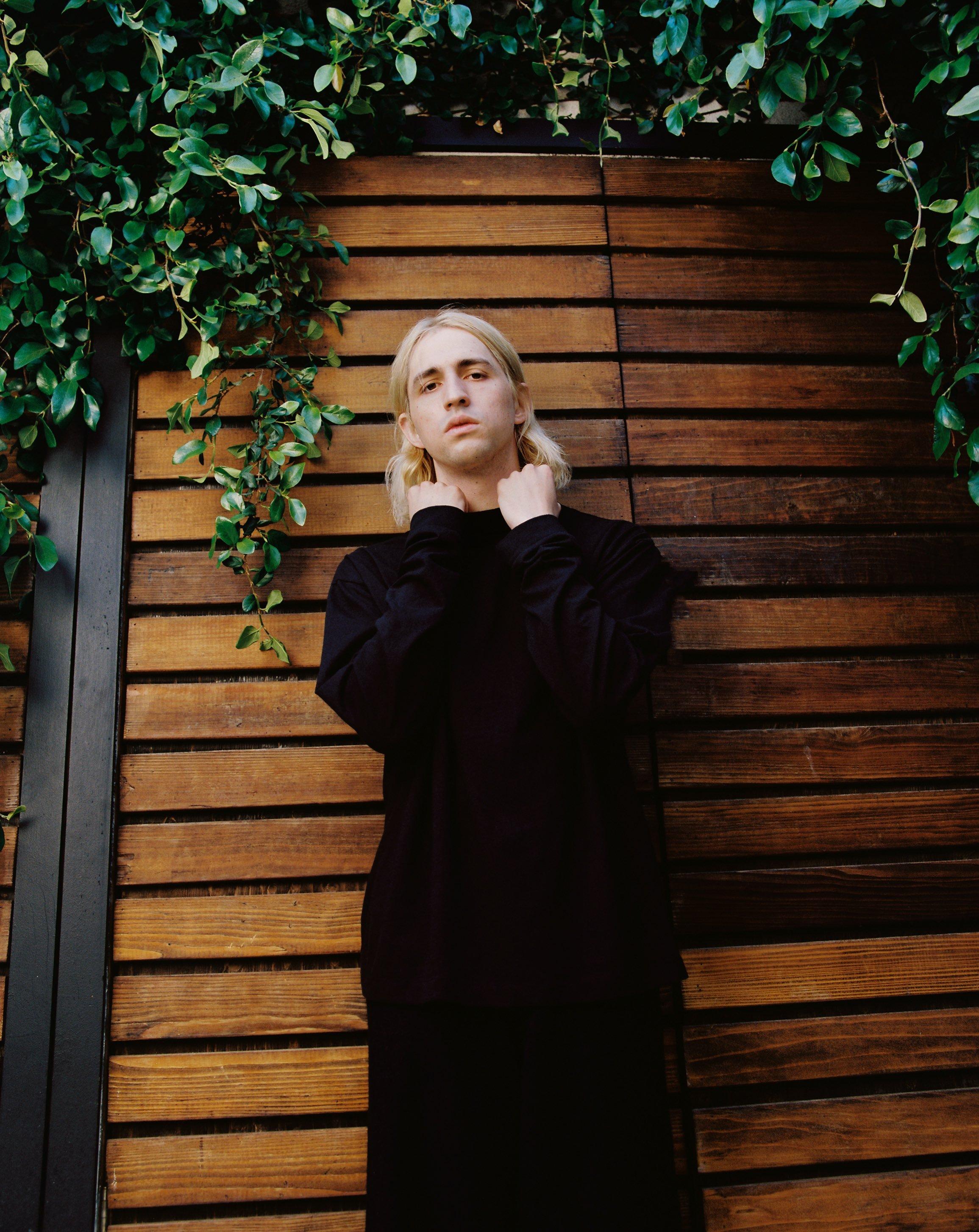 Porter Robinson poses in all-black, in front of a vine-covered fence