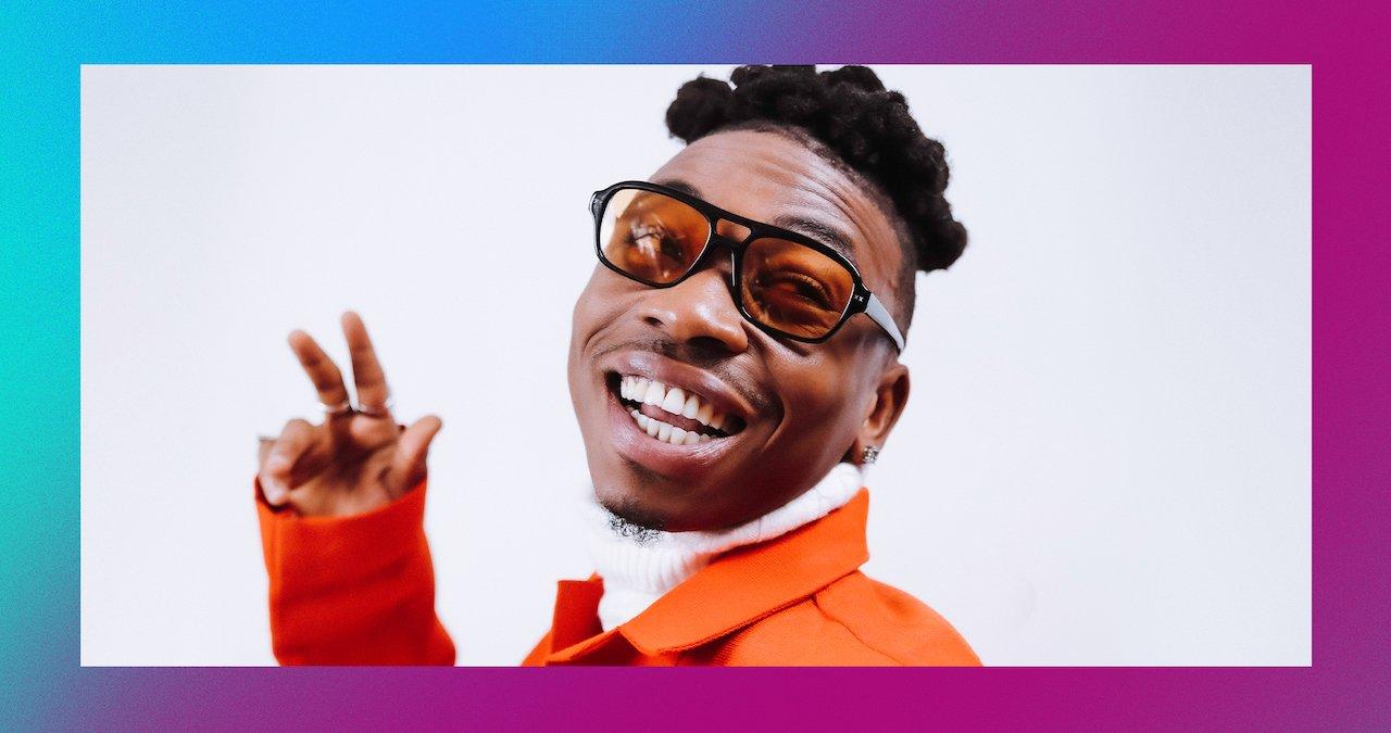 Mayorkun, wearing orange-tinted shades, looks over his shoulder and smiles