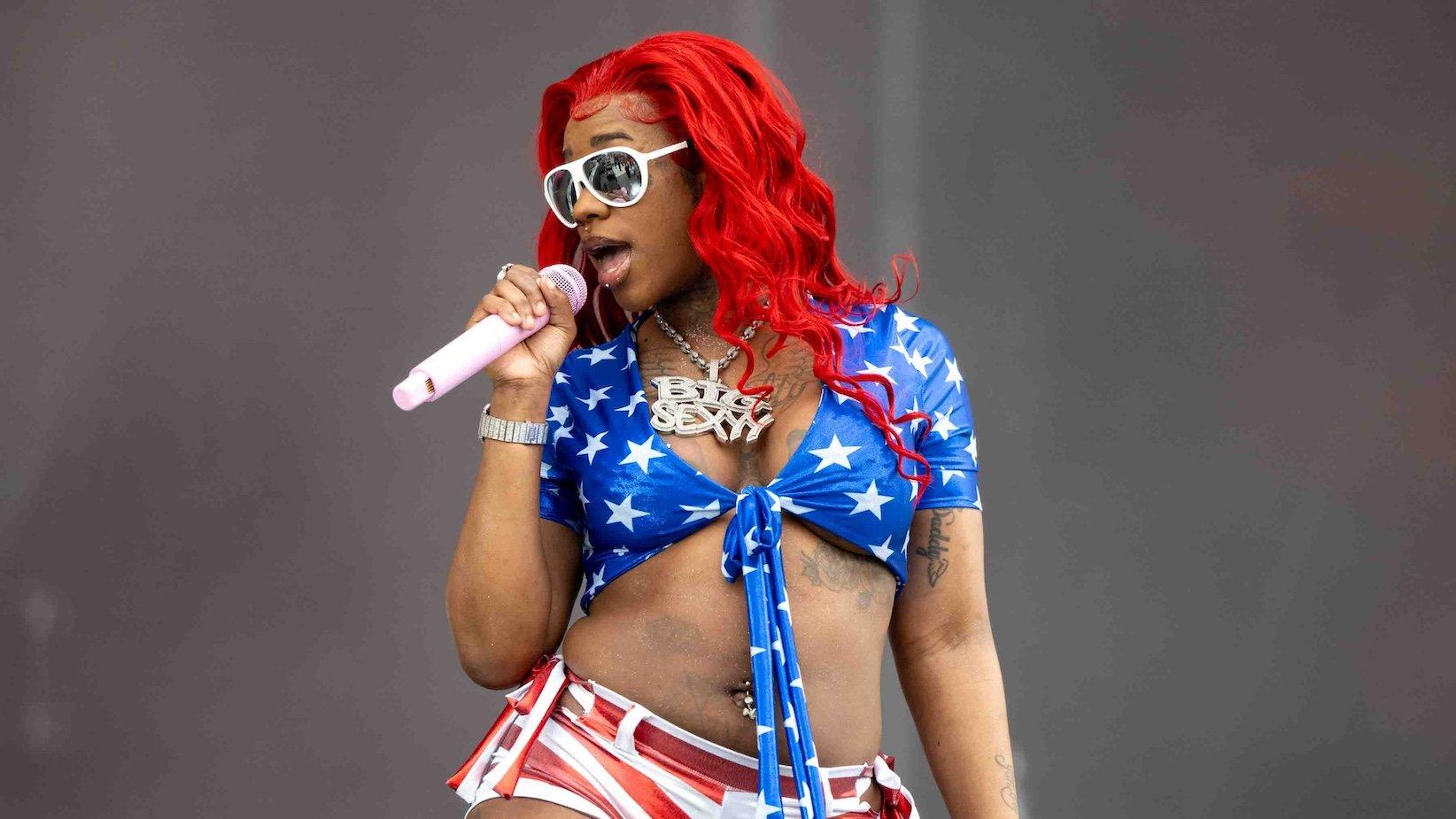 Photo of Sexyy Red performing onstage during at the 2024 Rolling Loud Festival in Los Angeles. She is wearing a blue bikini top with white stars, red and white shorts, white sunglasses, and bright red hair.