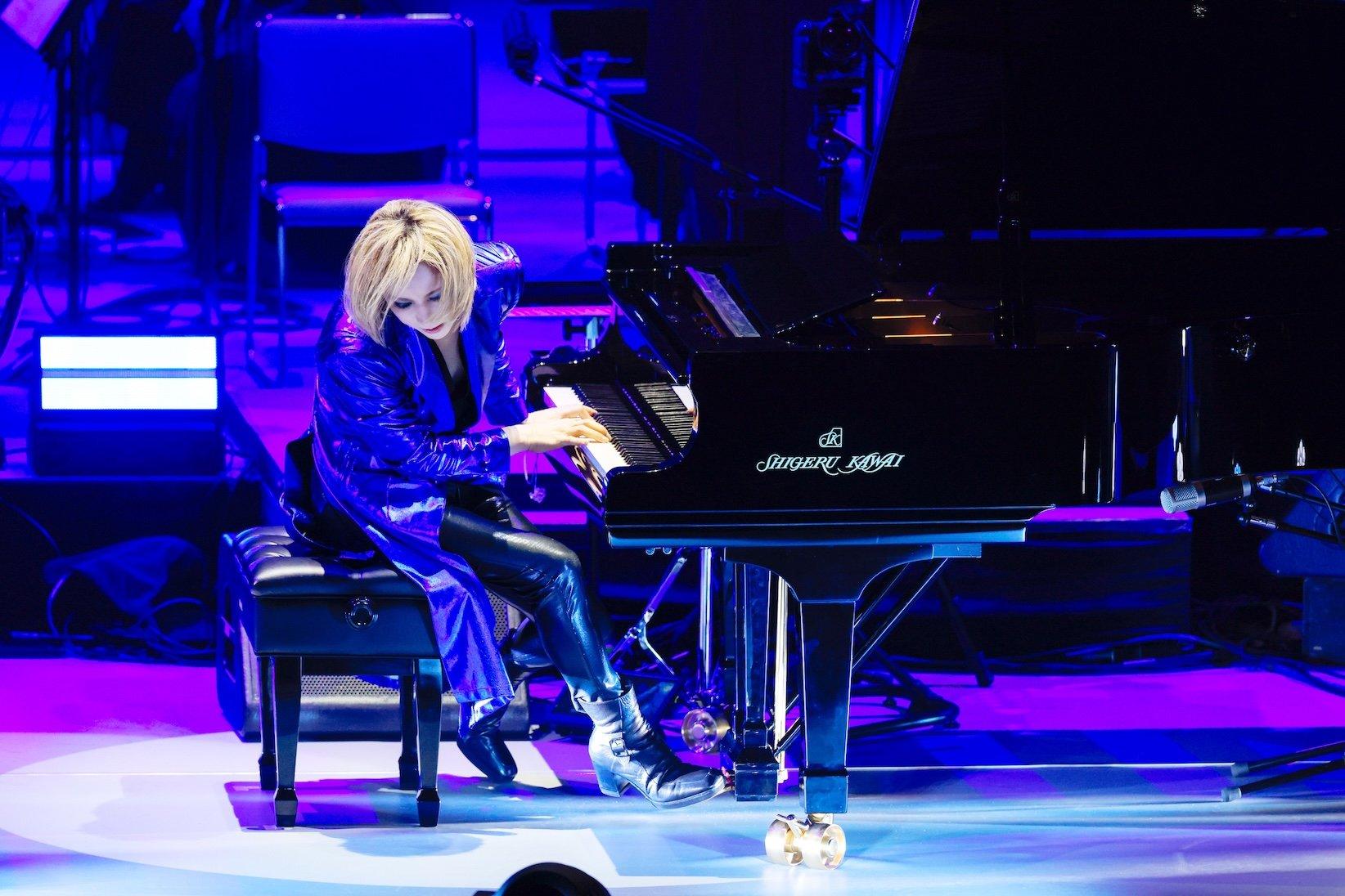 A photo of Yoshiki performing on a piano onstage. Yoshiki is wearing a shiny blue suit and black boots and performing on a black grand piano.